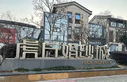 The sculptural entrance monument of Huqiu Wedding City, reading 虎丘婚纱城 / HUQIU BRIDAL CITY, with the villa-style designer shopfronts of the complex behind it.